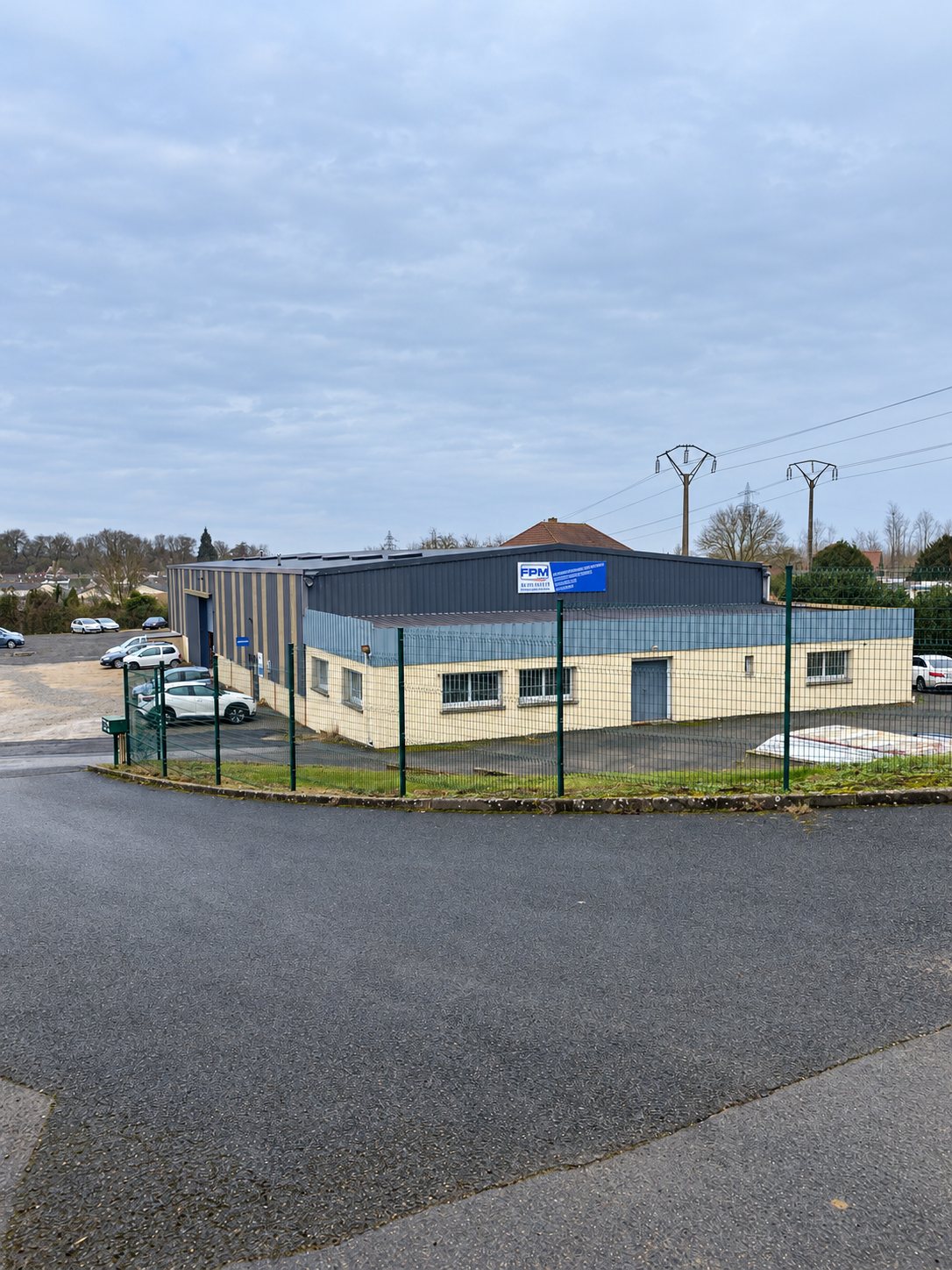 Garage Courmelles Soissons extérieur : un entrepôt aux façades beige et bleue portant l’enseigne FPM, entouré d’une clôture verte et de voitures garées, sous un ciel brumeux.