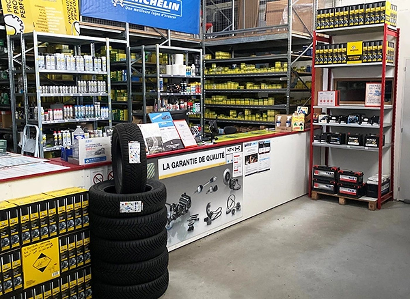 Intérieur d'un magasin de pièces automobiles avec un comptoir, une pile de pneus, des étagères d'huiles et de produits d'entretien et des batteries sur un rayonnage rouge.